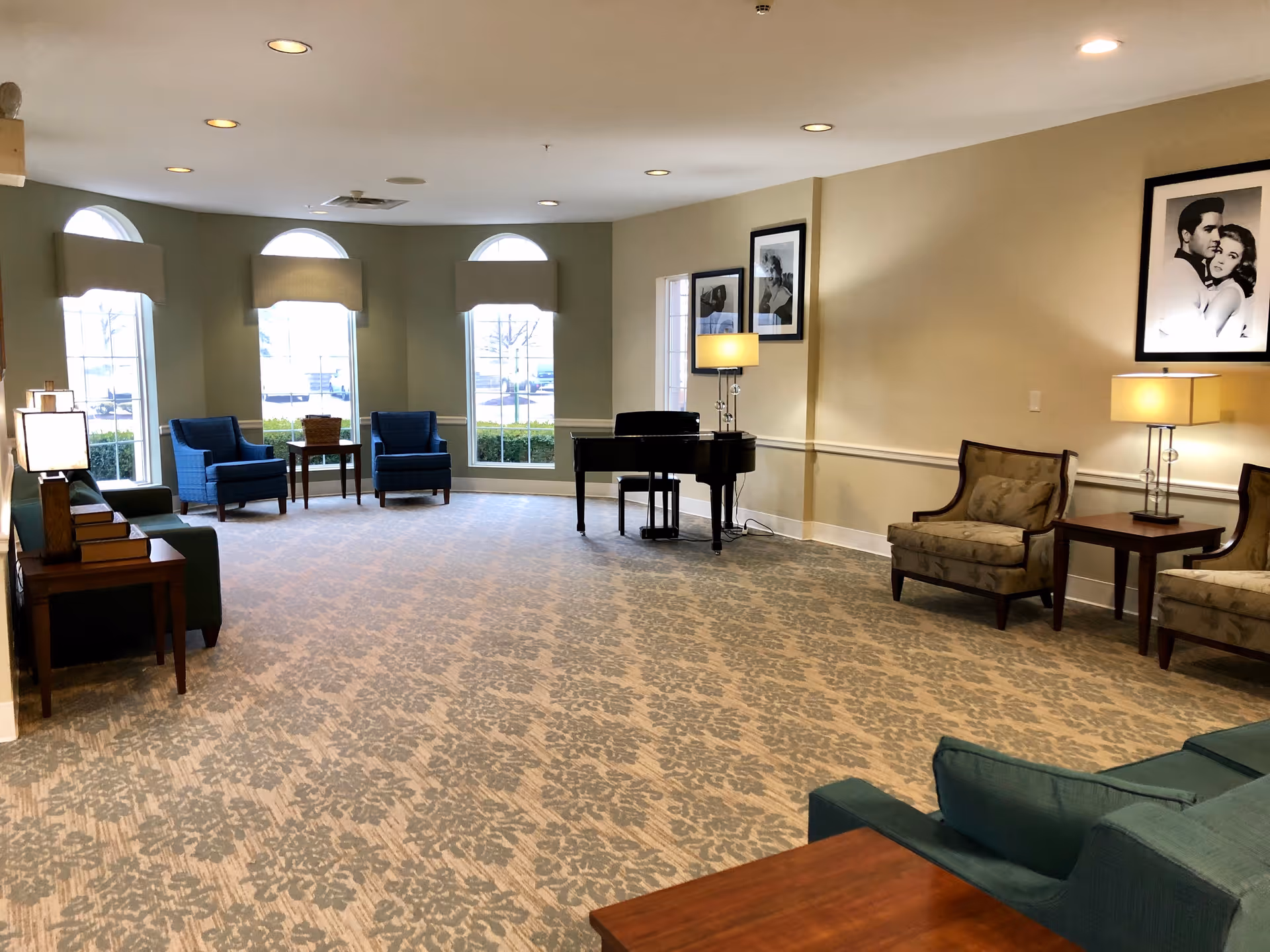 A spacious, well-lit living room with patterned carpet, several armchairs and sofas arranged around the room, a black grand piano near the wall, and framed black-and-white photos on the beige walls. Large windows with arched tops let in natural light.