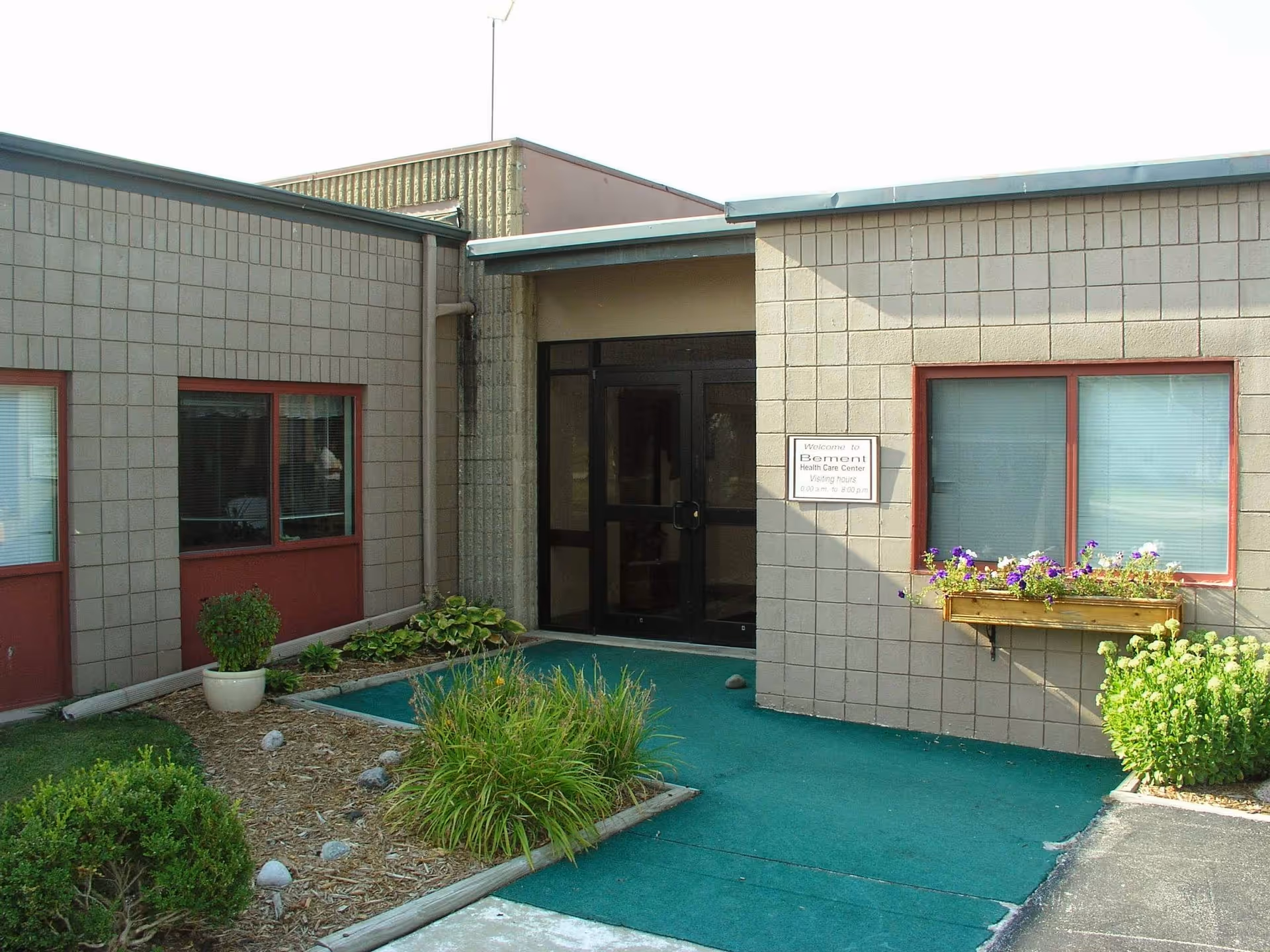 Exterior view of a single-story building with beige brick walls and red window frames. There is a glass double door entrance with a sign next to it that reads 'Welcome to Bement Health Care Center Visiting Hours 9:00 am to 8:00 pm'. The entrance is surrounded by small landscaped garden beds with green plants and flowers, and a green outdoor carpet leads to the door.