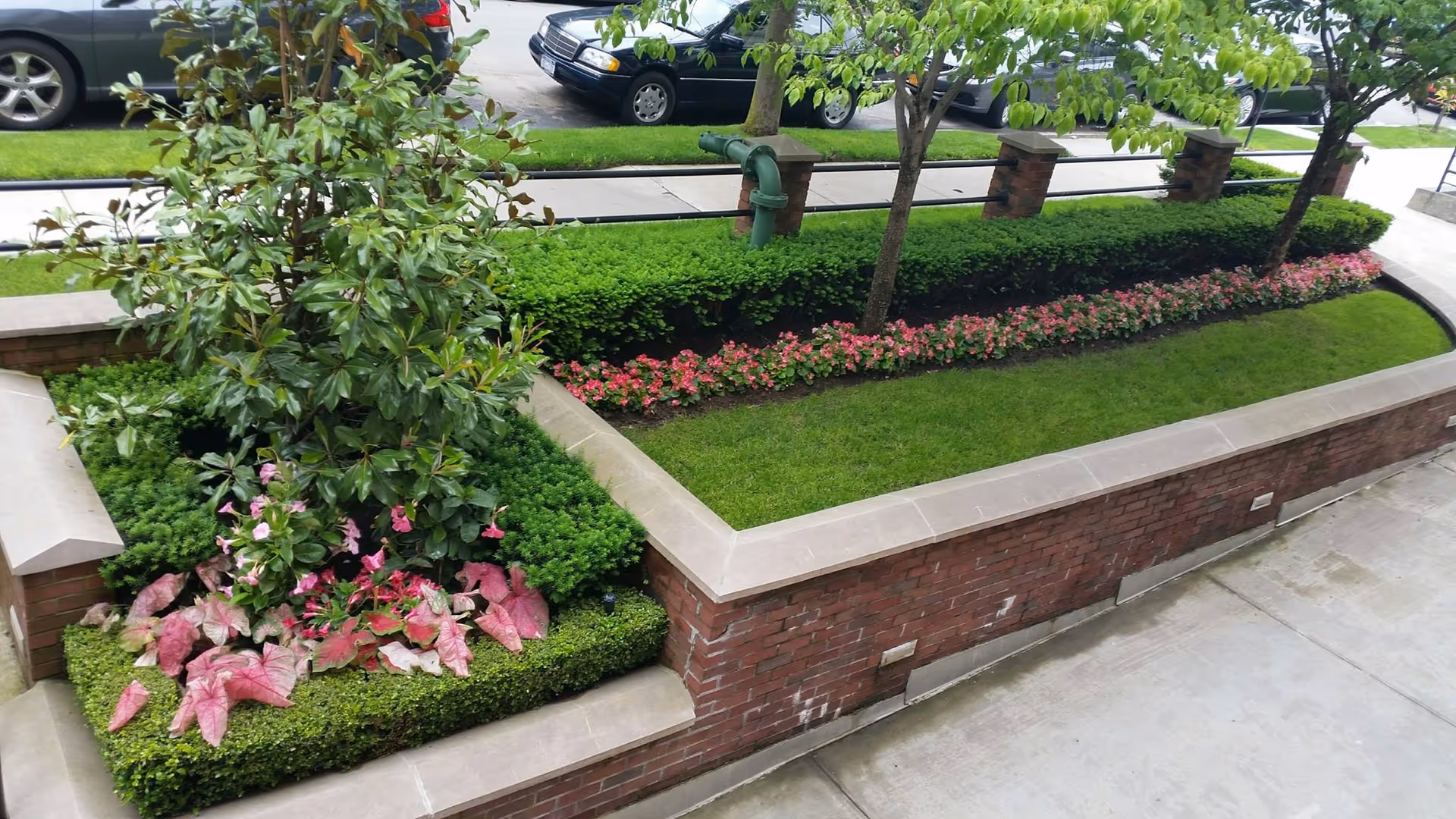 A landscaped outdoor garden area with neatly trimmed bushes, green grass, pink flowers, and small trees. The garden is bordered by a low brick wall with a stone cap. In the background, parked cars and a sidewalk are visible.