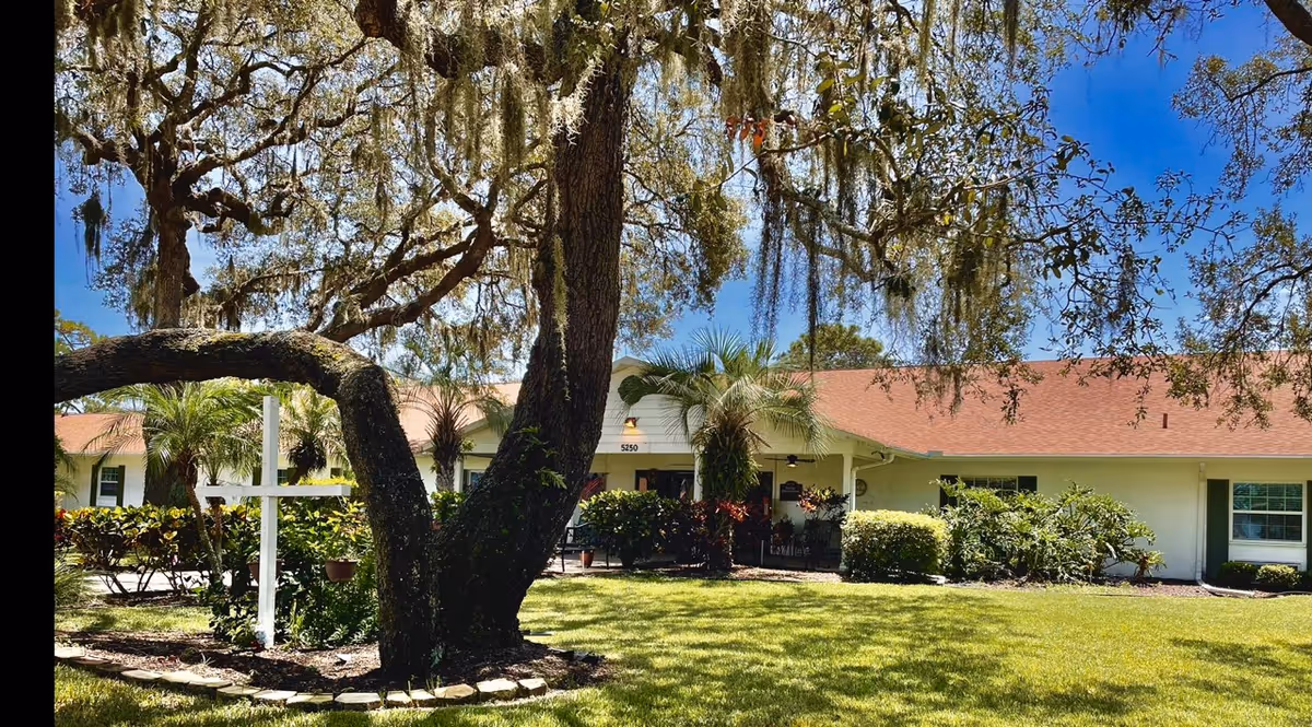Front exterior of a single-story light-colored building with a red roof, a large oak tree draped in Spanish moss, a white cross, and landscaped lawn and shrubs.