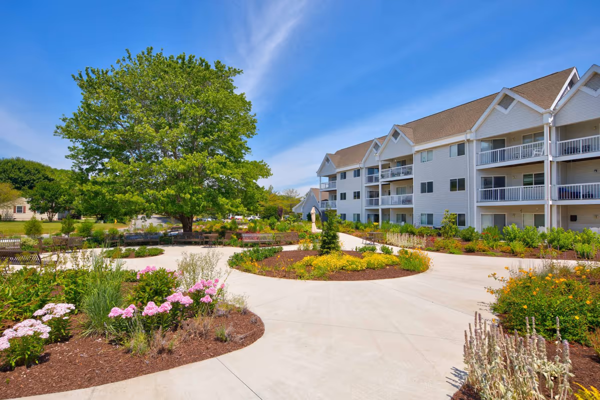 Outdoor garden area with a large tree, colorful flower beds, and multiple benches arranged along a curved concrete pathway. In the background, there is a three-story white residential building with balconies under a clear blue sky.