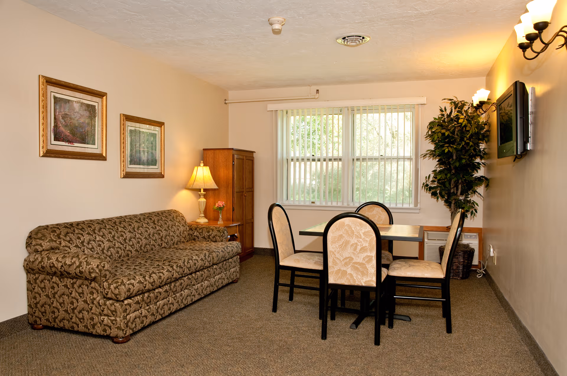 A cozy living room area with a patterned sofa on the left, a wooden cabinet with a table lamp and small flower vase next to it, a dining table with four upholstered chairs in front of a window with vertical blinds, a wall-mounted TV on the right wall, and a tall potted plant beside the TV.