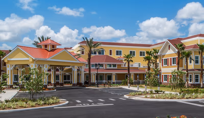 Exterior view of Buffalo Crossings Assisted Living facility showing a large yellow building with red roofs, palm trees, landscaped greenery, and a driveway with a covered entrance under a blue sky with scattered clouds.