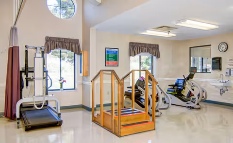 Bright rehabilitation/exercise room with a treadmill, stationary bikes, and a small wooden practice staircase near windows.