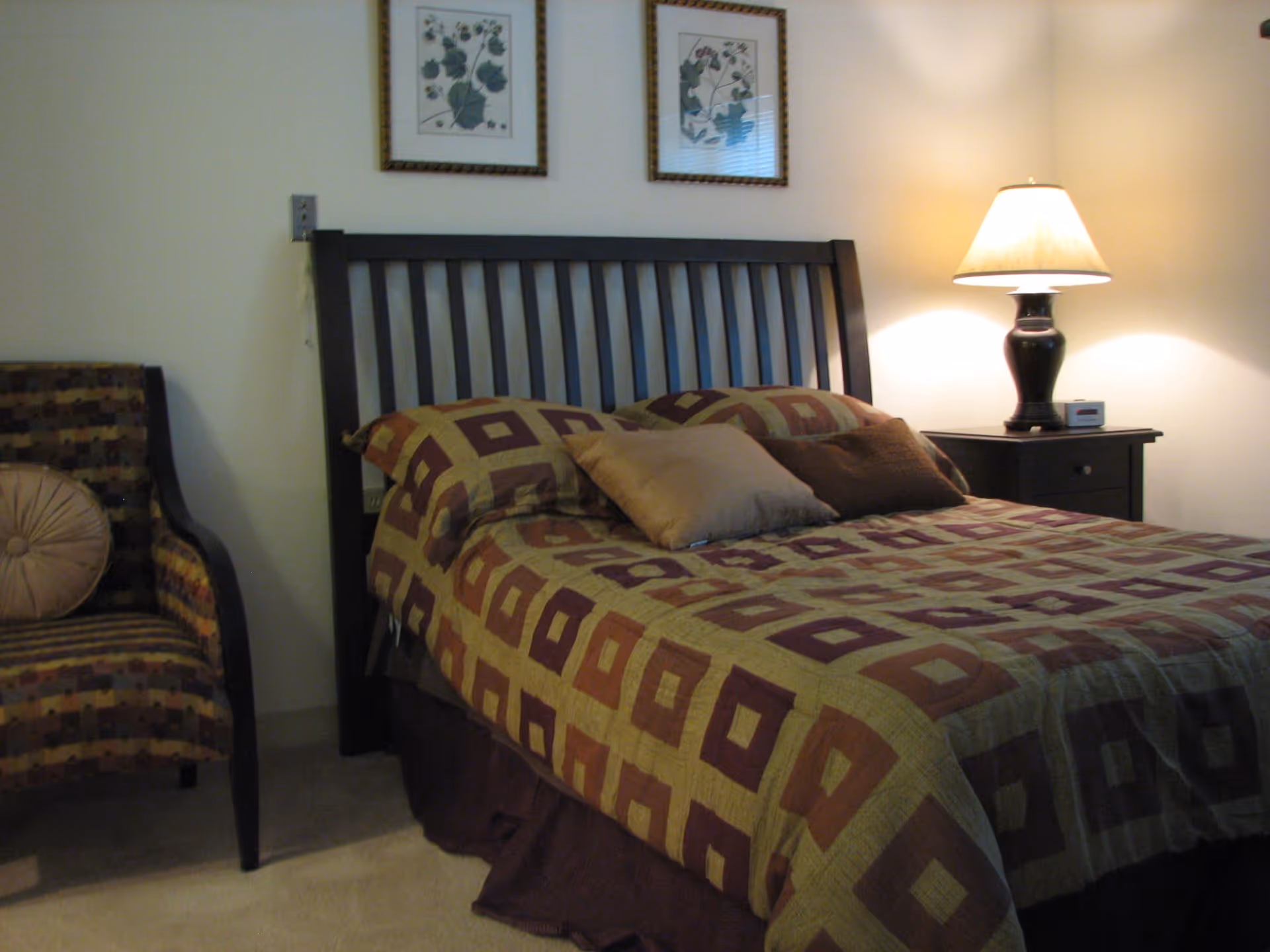 A bedroom with a bed featuring a patterned bedspread in shades of brown and green, two pillows, a wooden headboard, a nightstand with a lamp and a clock, and an upholstered chair with a round cushion.
