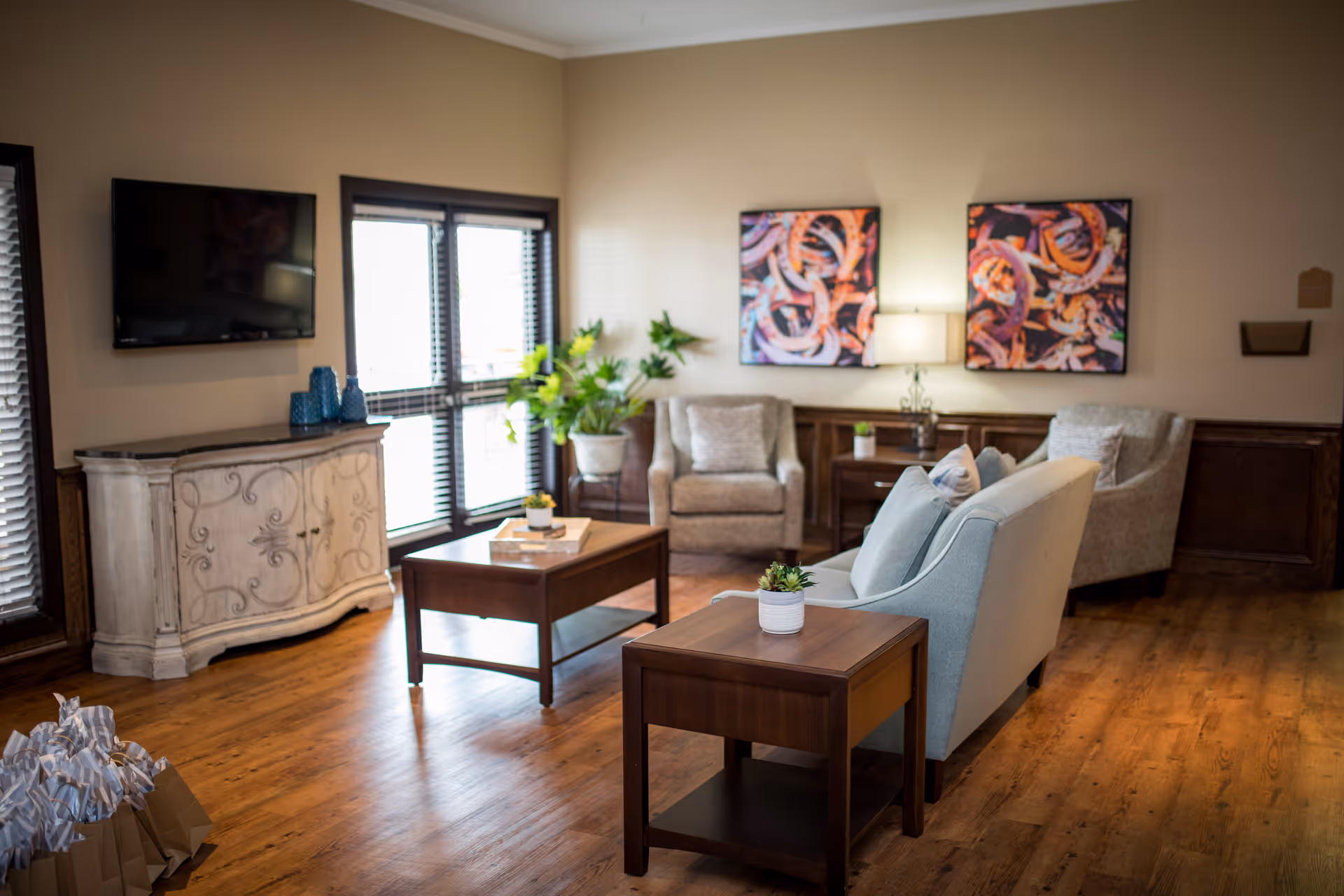 A cozy living room area with wooden flooring, a light blue sofa, two beige armchairs, a wooden coffee table, and a side table with small potted plants. There is a flat-screen TV mounted on the wall above a decorative cabinet, two abstract paintings on the wall, a table lamp, and a large potted plant near the window with blinds.