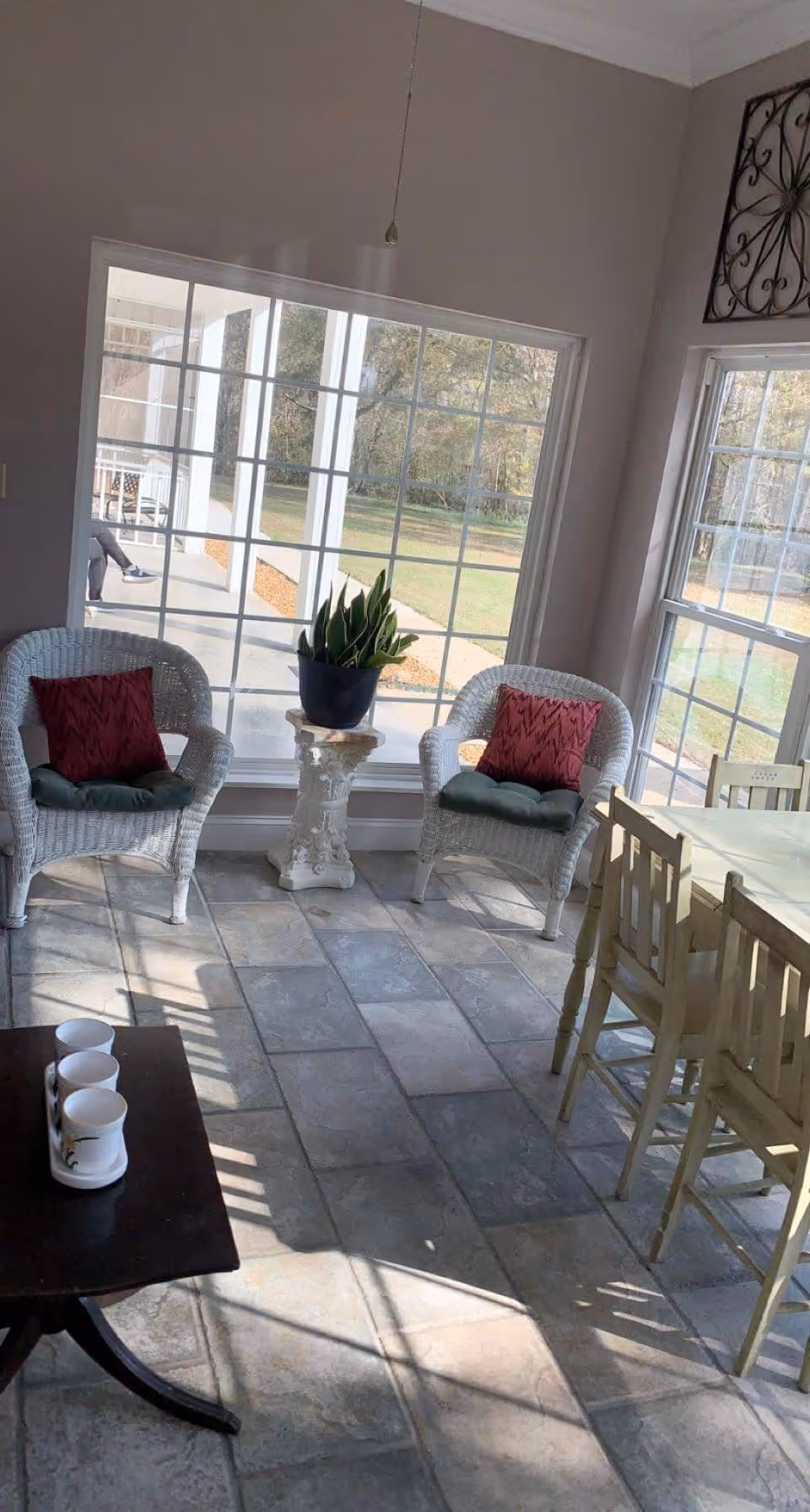 A sunlit sitting area with two white wicker chairs each with a red cushion and a green seat cushion, positioned around a small white pedestal table holding a potted plant. Large windows provide a view of an outdoor patio and grassy area. To the right, there is a wooden dining table with chairs. A small dark wooden table with three white mugs is in the foreground.