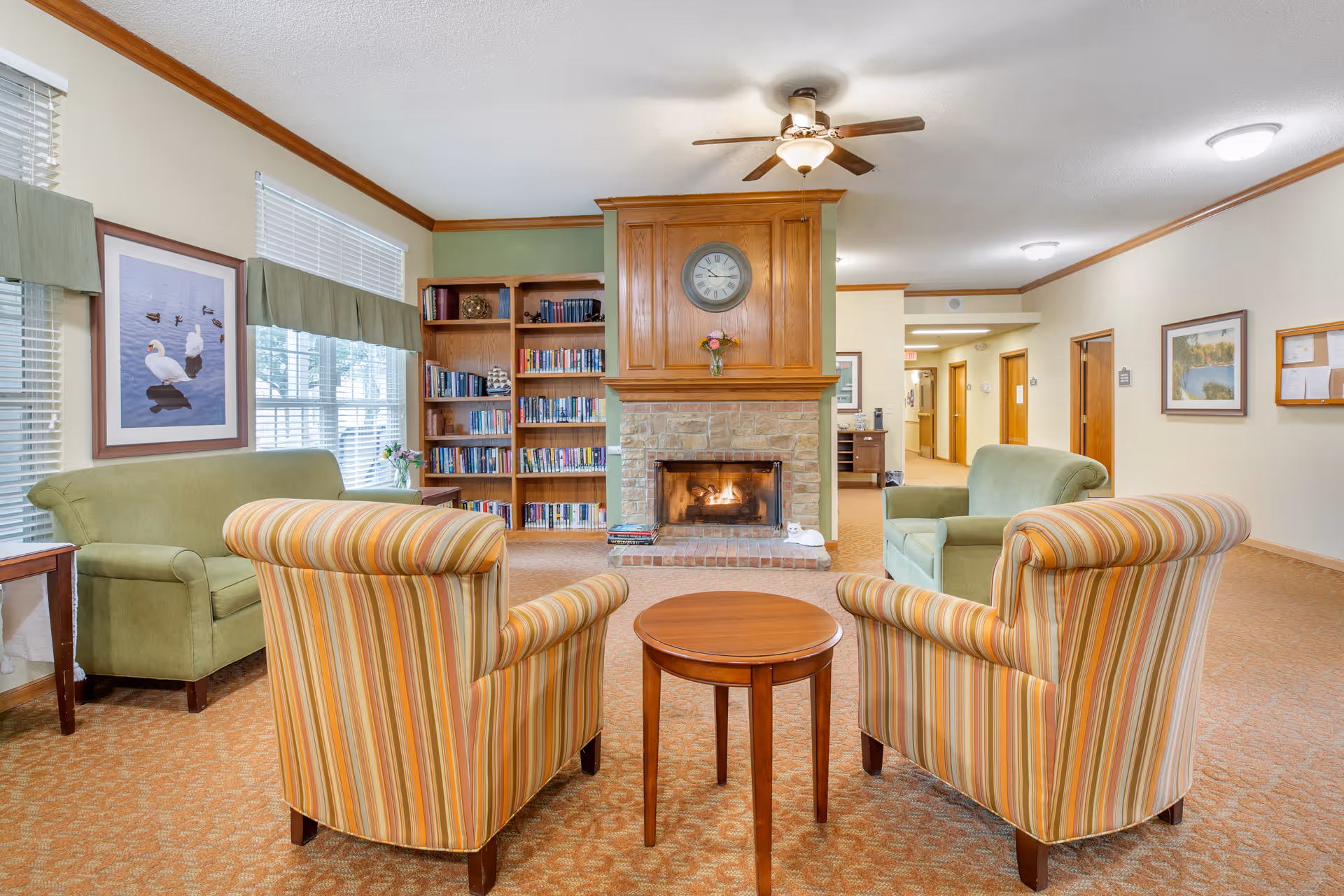 A cozy living room area in a senior living facility featuring a stone fireplace with a wooden mantle and a clock above it. In front of the fireplace are two striped armchairs with a small round wooden table between them. On either side of the fireplace are green upholstered sofas. To the left, there is a large window with blinds and a framed picture of swans on the wall. A wooden bookshelf filled with books is next to the fireplace. The room has beige walls with wooden trim and a ceiling fan with a light fixture.