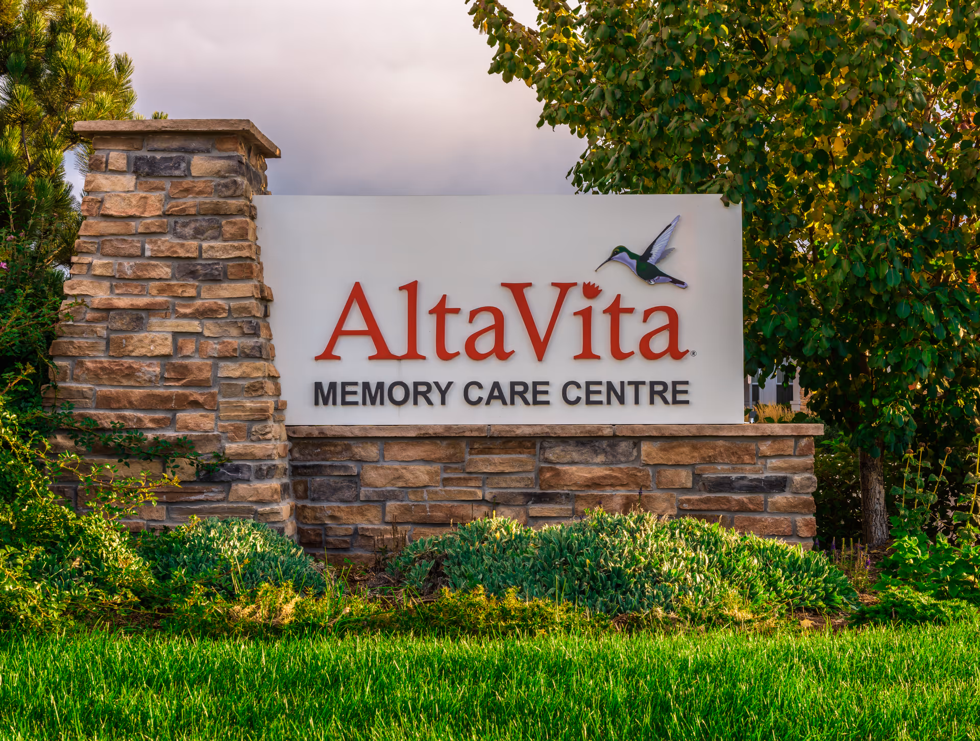 Stone and brick sign for AltaVita Memory Care Centre surrounded by green grass, bushes, and trees under a cloudy sky.