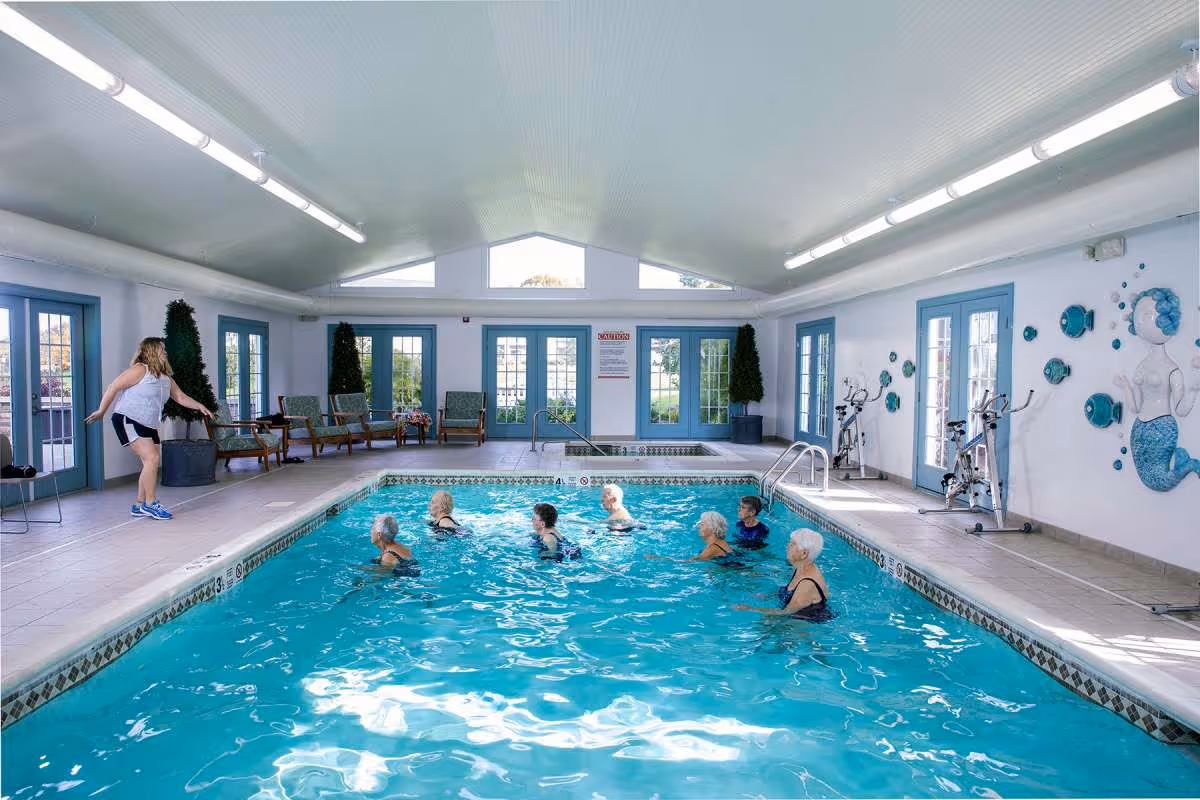 Indoor swimming pool with seven elderly women standing in the water and a younger woman on the poolside leading an exercise or activity. The room has large windows and glass doors with blue frames, several chairs along the walls, two stationary bikes, and a wall decoration of a mermaid.