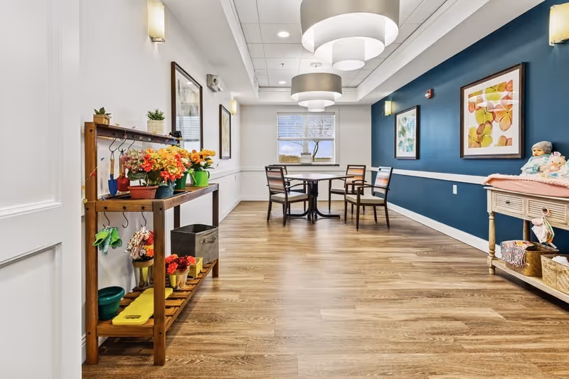 Bright communal dining/activity room with a wooden shelf of potted flowers in the foreground, a round table and chairs under pendant lights, and a teal accent wall with artwork.