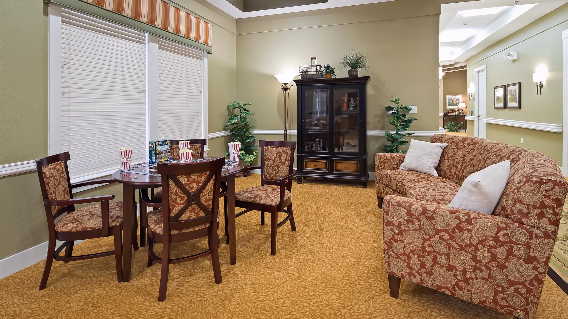 A cozy common area in an assisted living facility featuring a round wooden table with four upholstered chairs, each with a red and gold floral pattern. On the table are four cups with red and white stripes. To the right is a matching upholstered sofa with two white pillows. The room has beige walls, a patterned carpet, two potted plants, a tall floor lamp, and a dark wooden cabinet with glass doors. There are two large windows with closed white blinds and a striped valance above them.