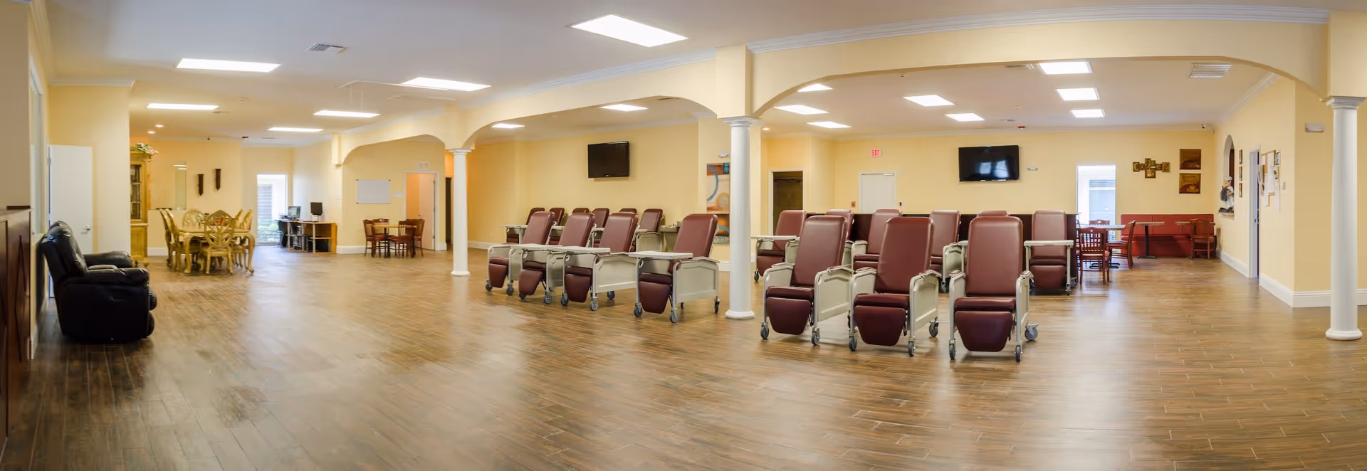 A spacious adult daycare facility room with wooden flooring and beige walls. The room contains multiple reclining chairs with wheels arranged in rows, several dining tables with chairs, a black recliner chair, two wall-mounted televisions, and a few computer desks in the background. The room is well-lit with ceiling lights and has white columns supporting the ceiling.