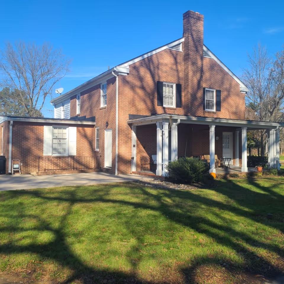 A two-story brick house with white trim and black shutters, featuring a covered porch with chairs and a small table. The house is surrounded by a green lawn and leafless trees under a clear blue sky.