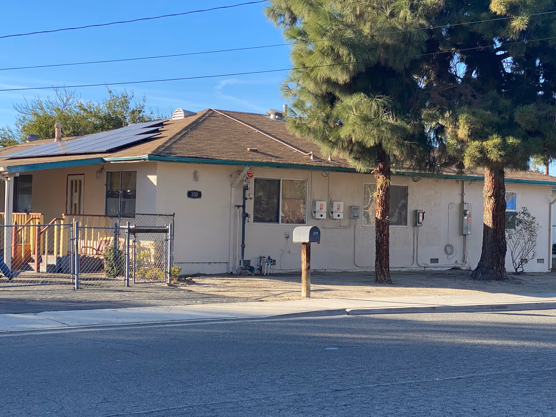 Single-story beige building with a porch, chain-link fence, mailbox, and large pine trees along the front.