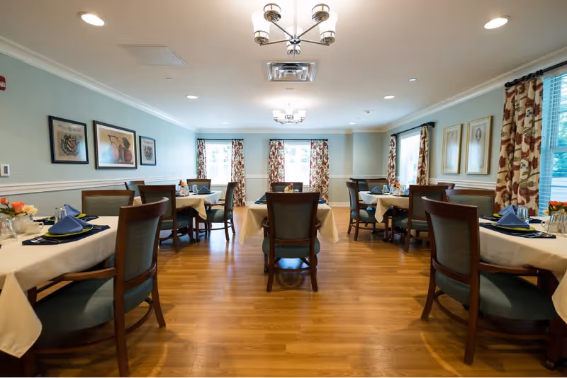 Bright dining room with several neatly set tables, wooden chairs, and patterned curtains.