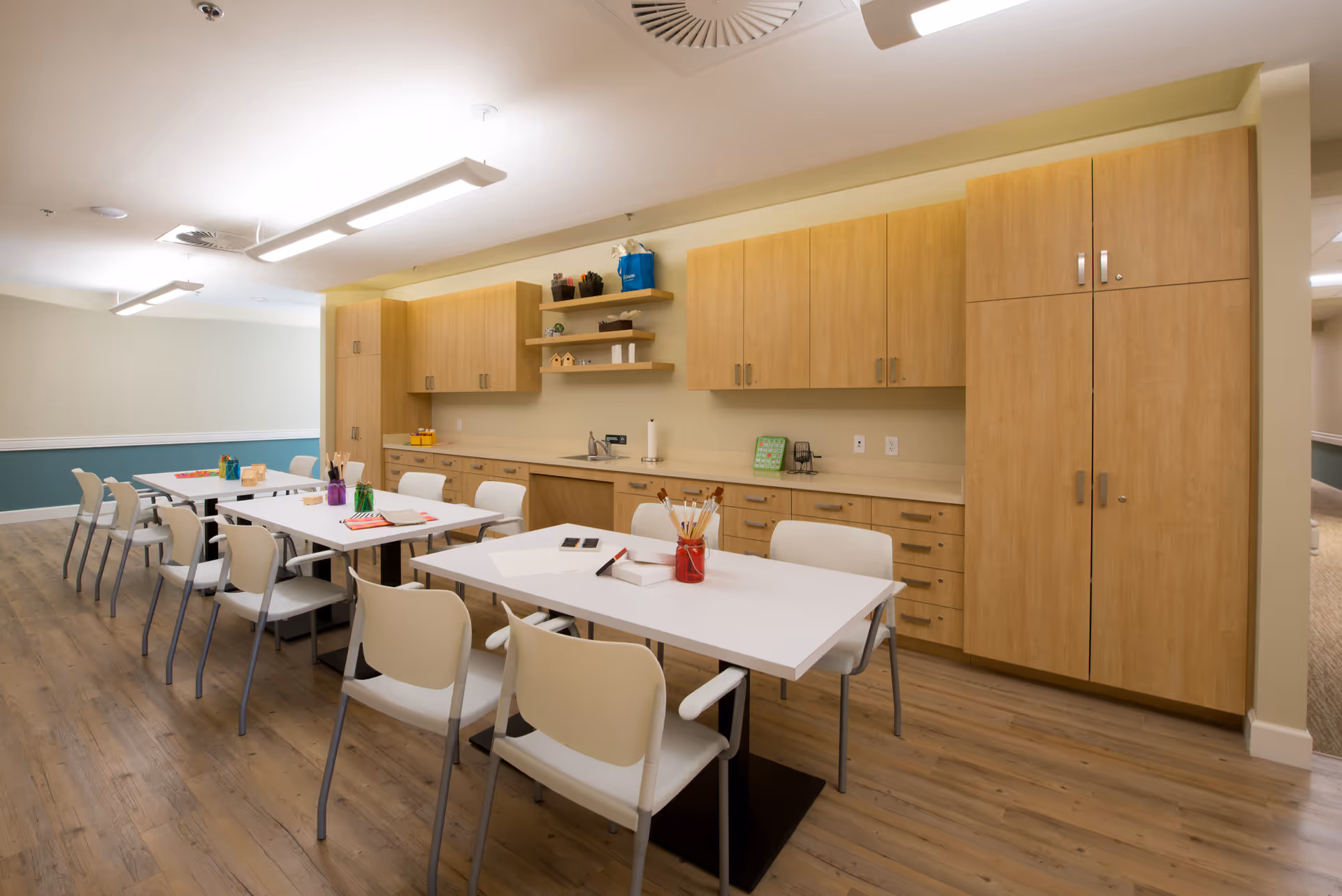 A well-lit activity room with white tables and beige chairs arranged in rows. The tables have various art supplies such as paintbrushes, colored pencils, and paper. The back wall features light wood cabinets and shelves with small decorative items. The floor is wooden, and the walls are painted in a light beige and teal color combination.