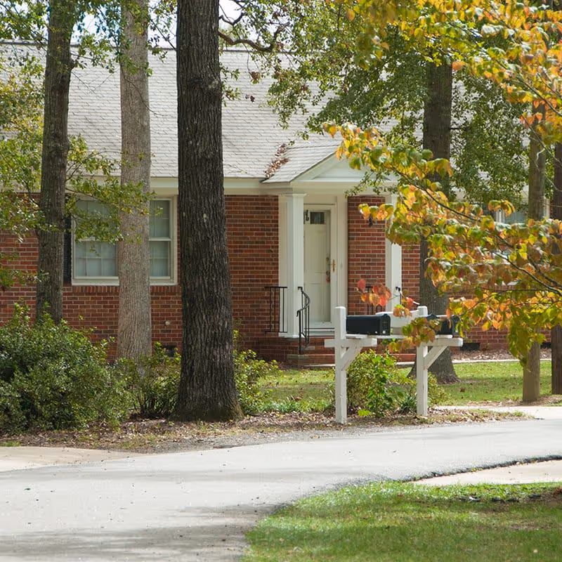 Exterior view of a brick building with a white door and windows, surrounded by trees and greenery. There are two black mailboxes mounted on a white wooden post near the sidewalk.