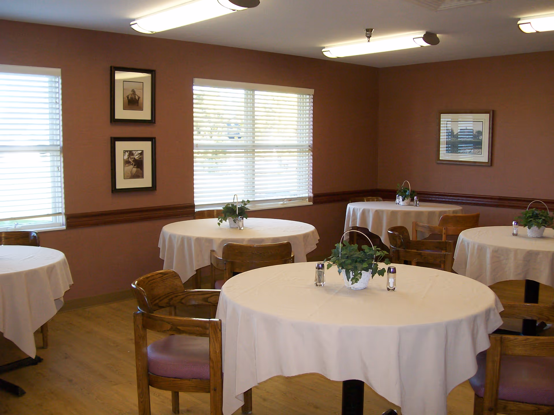 Small dining room with round tables covered in white tablecloths, wooden chairs, and potted centerpieces.