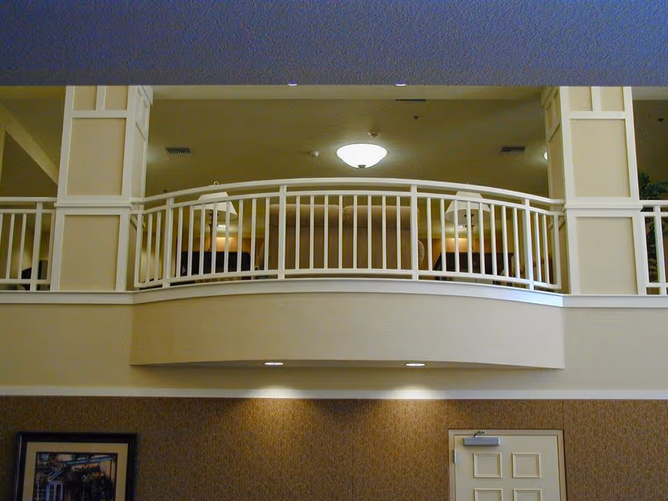 Interior view of a senior living facility showing a second-floor balcony with white railings overlooking a lower level. The upper level has beige walls, a ceiling light fixture, and lamps on side tables. Below, there is a closed door and a framed picture on the wall.