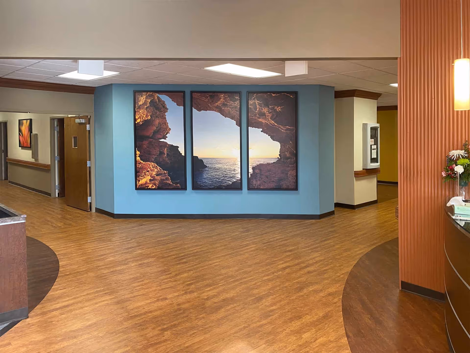 Interior hallway of a healthcare facility with wood-patterned flooring and a blue accent wall featuring a three-panel artwork of a rocky cave opening to a sunset over the ocean. There are doors and framed pictures along the hallway, and a reception desk with flowers on the right side.
