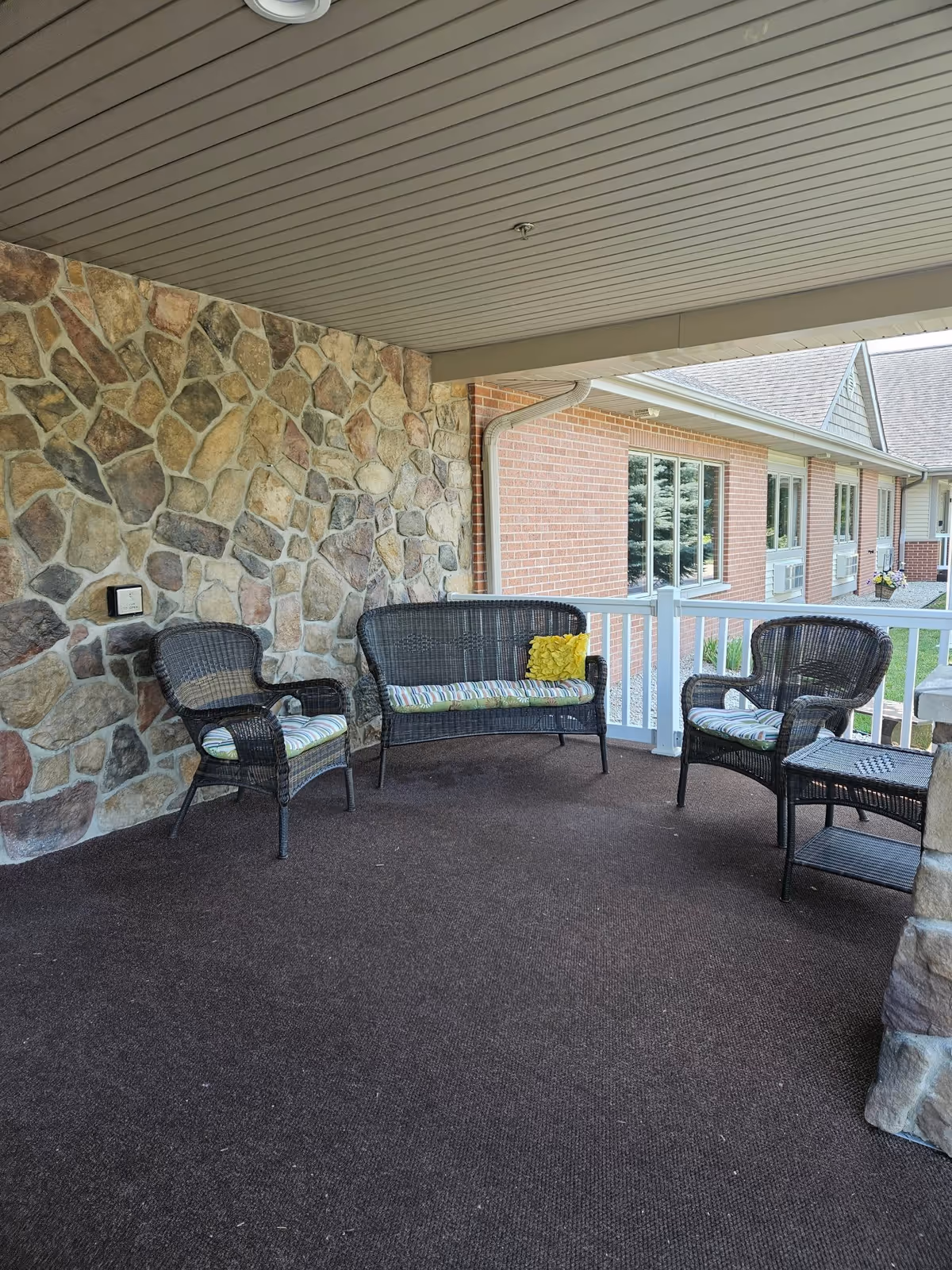Covered outdoor patio area with stone and brick walls, featuring three wicker chairs and a wicker loveseat with striped cushions and a yellow pillow, overlooking a grassy area and building windows.