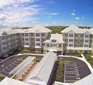 Aerial view of a large, multi-story independent living community building with white and beige exterior walls and multiple windows. The building surrounds a courtyard area with a covered walkway and landscaped greenery. There is a parking lot with marked spaces, including handicapped spots, adjacent to the building. The sky is partly cloudy with blue patches visible.