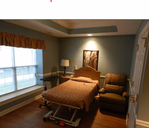 A small bedroom with an adjustable hospital-style bed, a recliner, a bedside lamp, and a window.