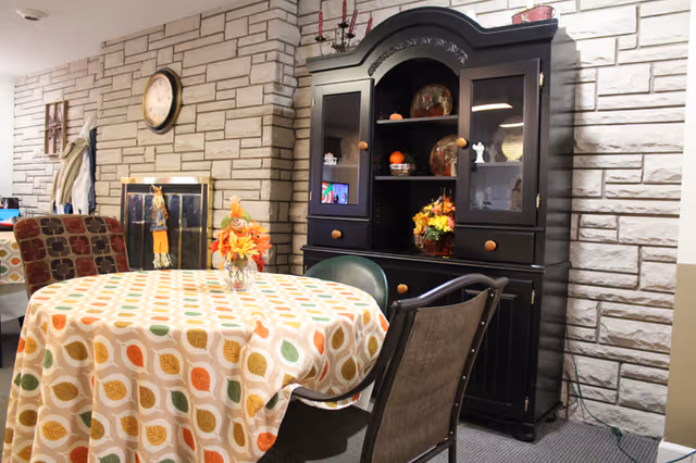 A cozy interior room with a round table covered by a colorful leaf-patterned tablecloth, surrounded by various chairs. Behind the table is a black wooden cabinet with glass doors displaying decorative items and autumn-themed decorations. The walls are made of light-colored stone bricks, and a clock is mounted on the wall above a fireplace with a scarecrow decoration nearby.