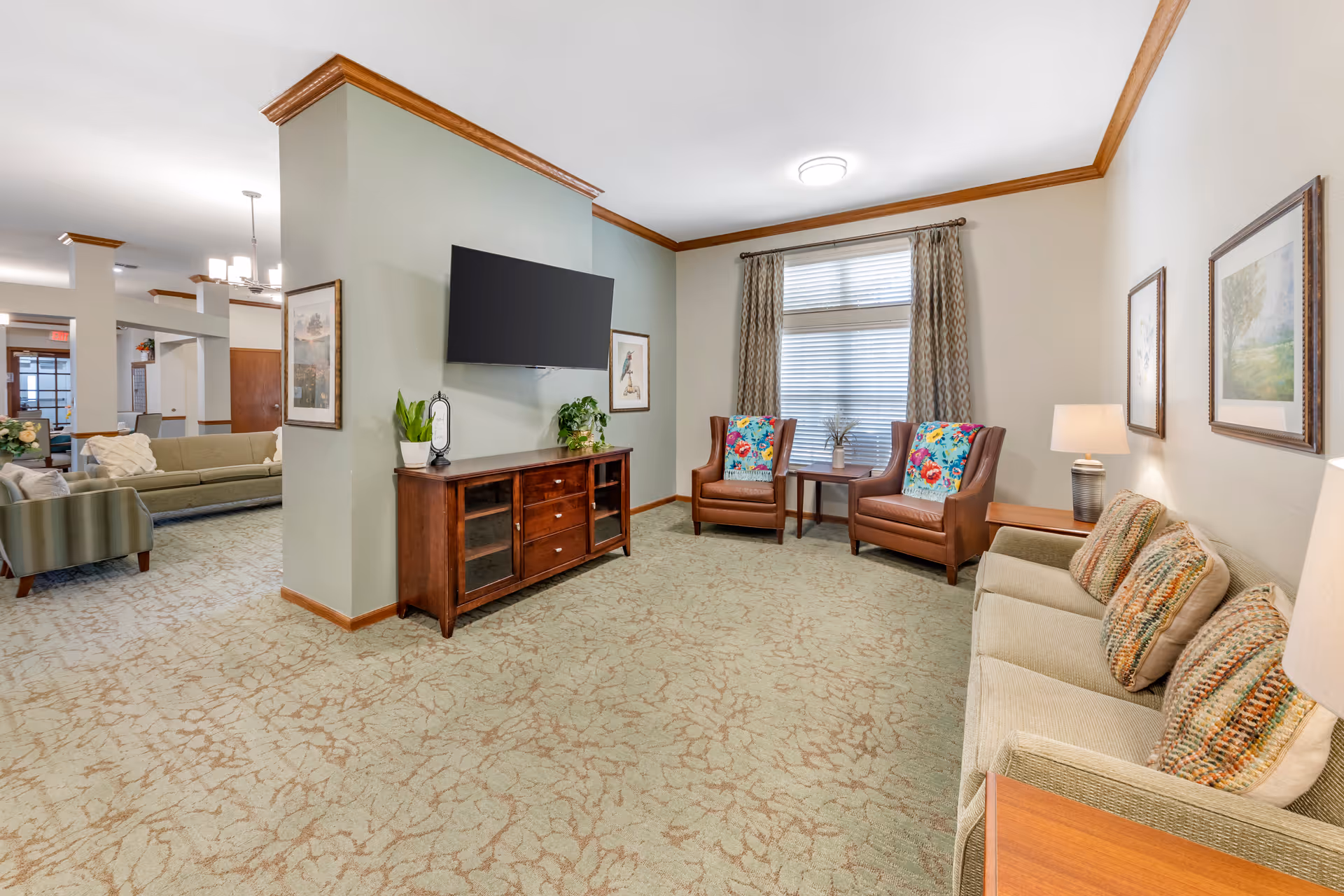 Well-lit communal living room with sofas, armchairs, a wall-mounted TV on a partial partition, and a window with curtains.