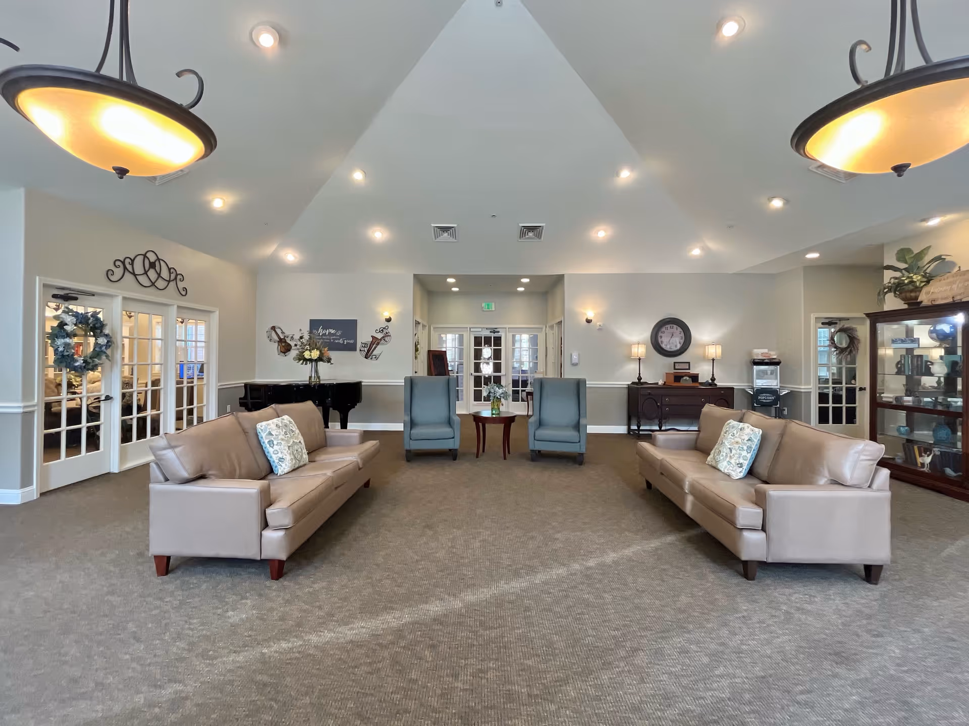 A spacious and well-lit living room area in Creekside Alzheimer's Special Care Center featuring two beige leather sofas with patterned pillows facing each other, two blue armchairs with a small wooden table between them, a black grand piano against the back wall, decorative wall art, a clock above a wooden sideboard with lamps, and glass doors on either side leading to other rooms. The ceiling is vaulted with recessed lighting and two hanging light fixtures.