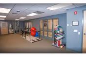 Interior view of a rehabilitation center room with exercise equipment including parallel bars, a red exercise ball, and various therapy tools organized on a rack against a blue wall.