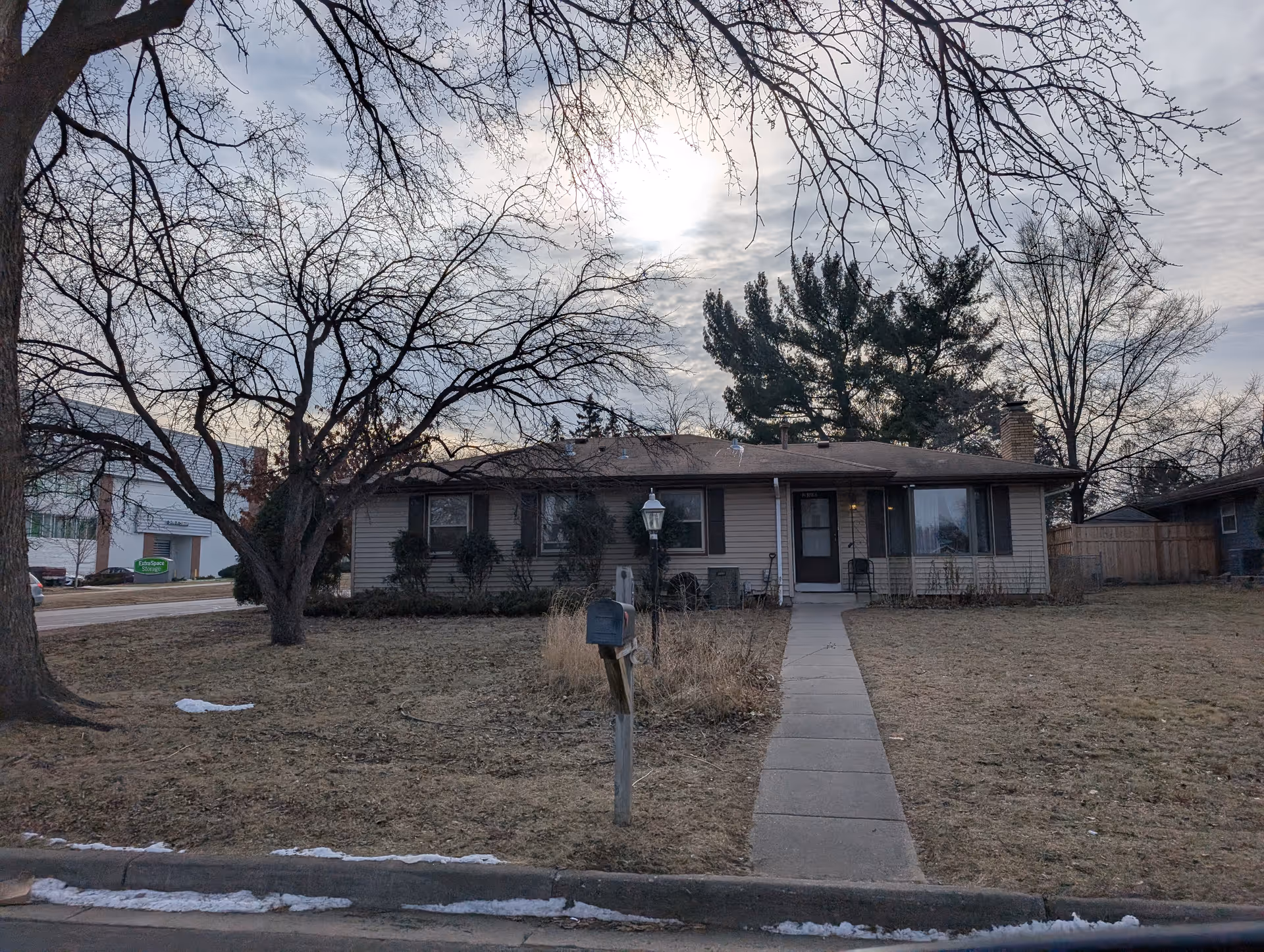 Single-story residential building with a front walkway leading to the entrance, surrounded by leafless trees and dry grass under a cloudy sky.