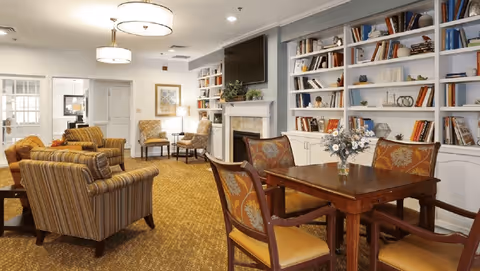Comfortable communal living room with upholstered chairs, a table, fireplace, built-in bookshelves and a wall-mounted TV.