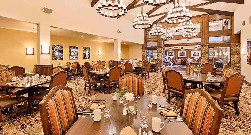 A spacious dining room with multiple wooden tables and upholstered chairs arranged neatly. Each table is set with glasses, mugs, napkins, and silverware. The room features warm lighting with large chandeliers hanging from a high ceiling with exposed wooden beams. The walls are decorated with framed pictures and there is a carpeted floor with a patterned design. In the background, there is a seating area with people conversing.