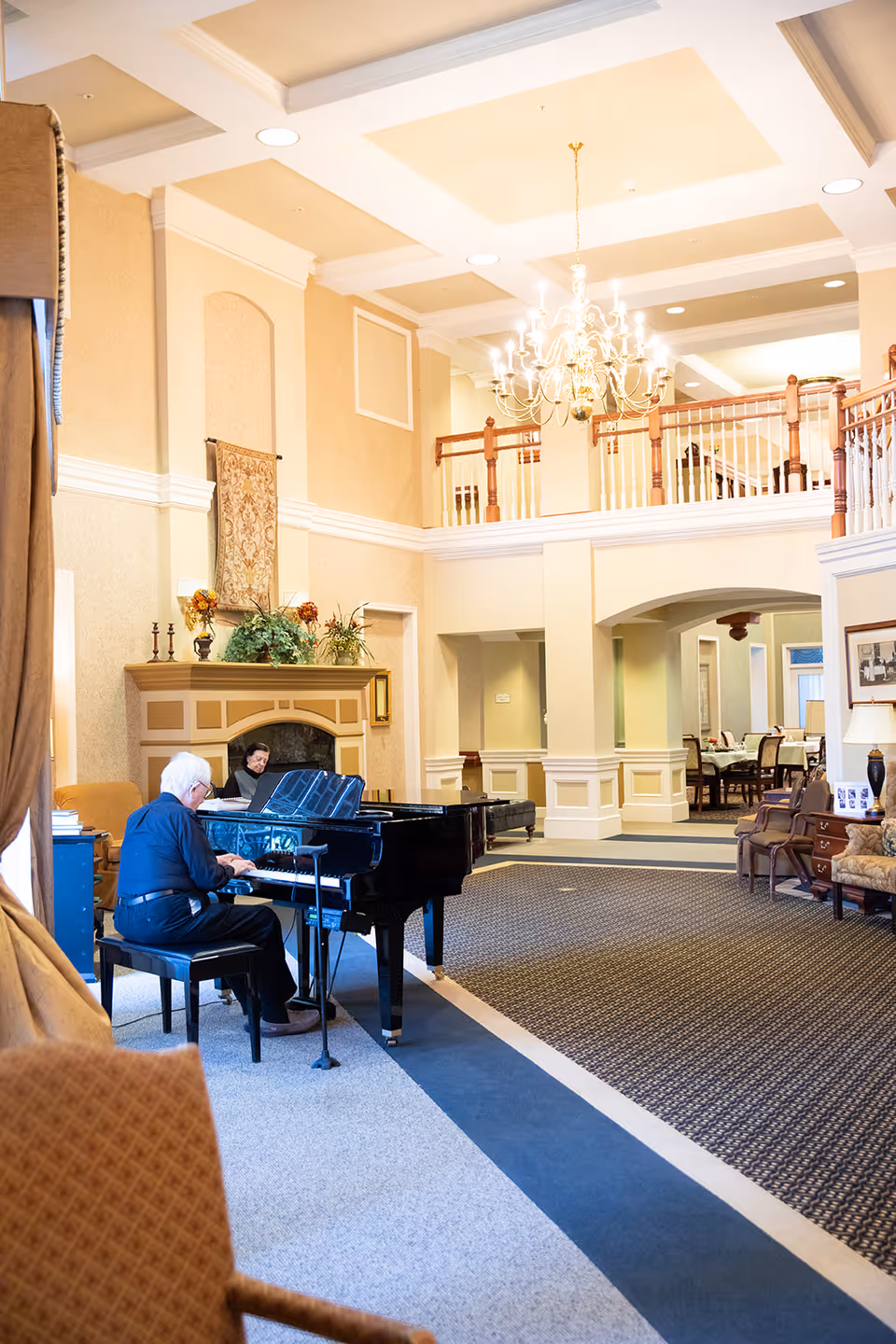 A spacious and elegant common area in a senior living facility with high ceilings, a chandelier, and a grand piano being played by an elderly man. Another person is seated nearby, and the room features comfortable seating, a fireplace, and decorative plants.