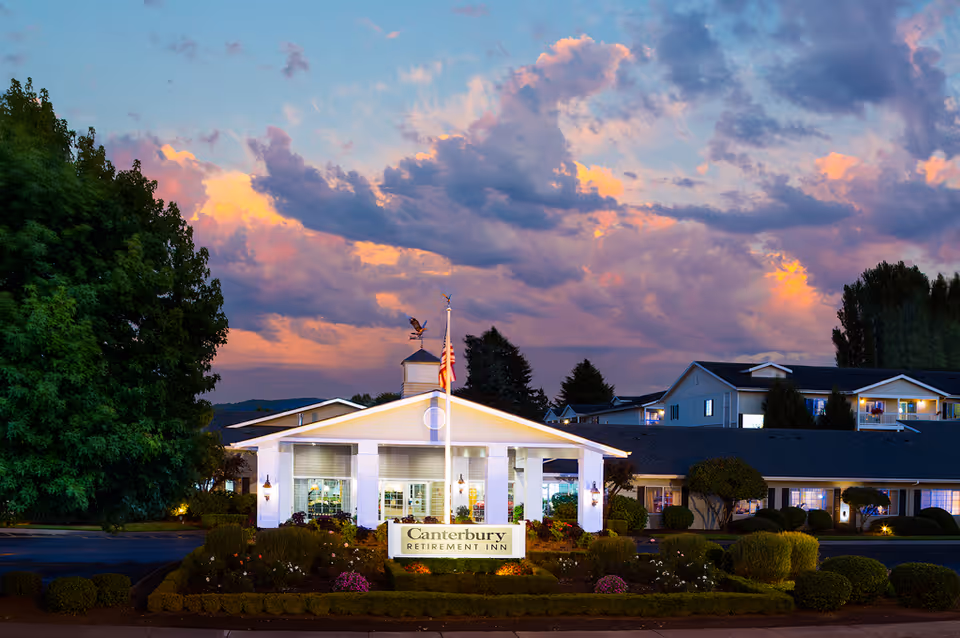 Exterior view of Canterbury Retirement Inn building at dusk with a colorful sky and clouds in the background, well-maintained landscaping, and an American flag on a flagpole in front.