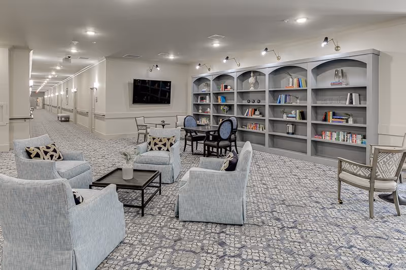 Spacious senior living common area with upholstered chairs, tables, built-in bookshelves and a wall-mounted TV along a long carpeted corridor.