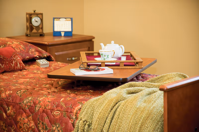 A cozy bedside scene with a patterned red bedspread, a green throw, an overbed tray holding a teapot and cup, and a nightstand with a clock.