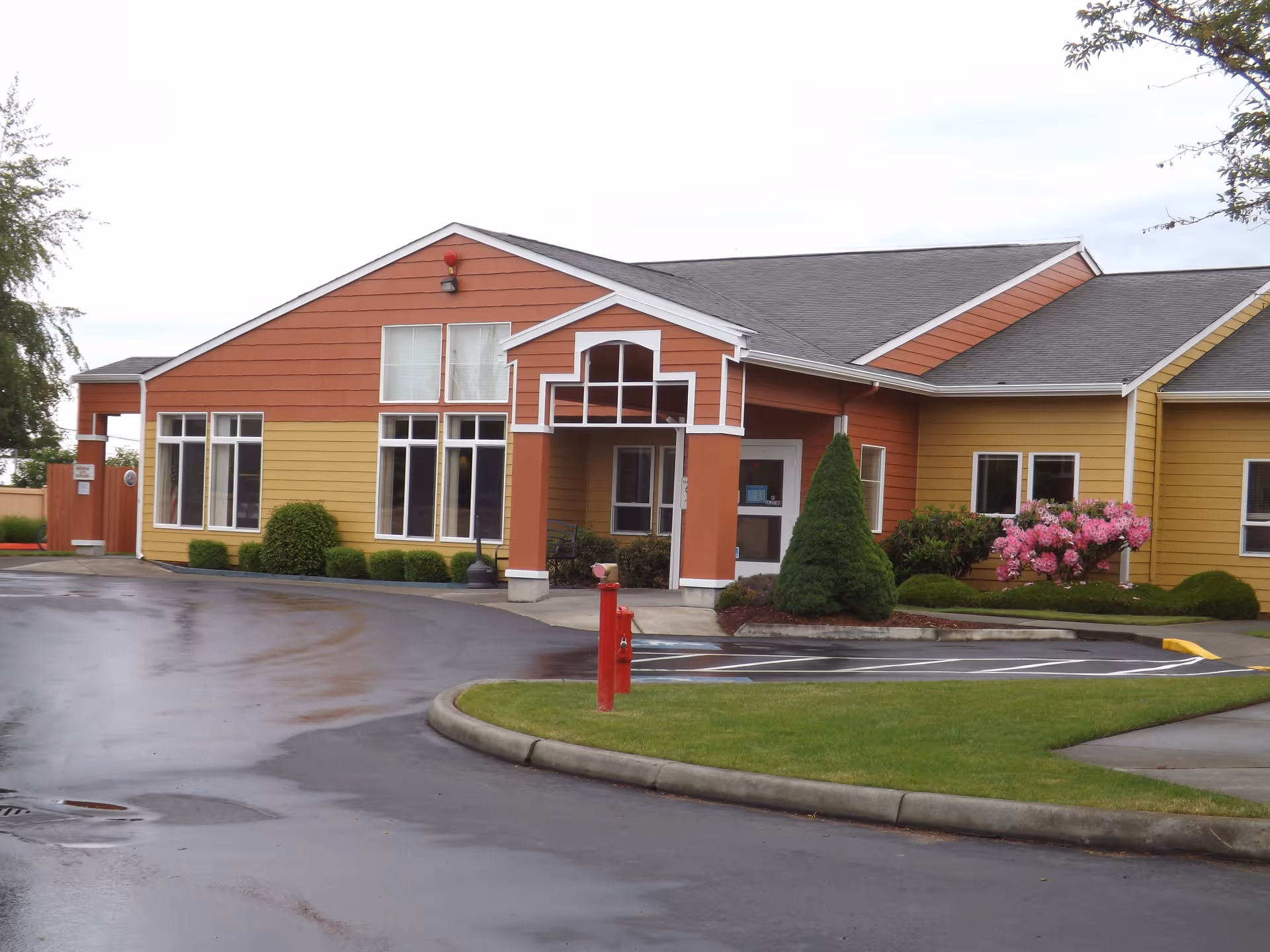 Front entrance of a low-rise retirement community building with orange and yellow siding, landscaped shrubs and a circular driveway on wet pavement.