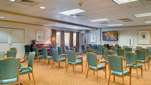 A spacious room with multiple rows of green cushioned chairs arranged facing a television mounted on the wall. The room has beige walls, a patterned carpet, framed artwork, an American flag, and windows with brown curtains allowing natural light in.