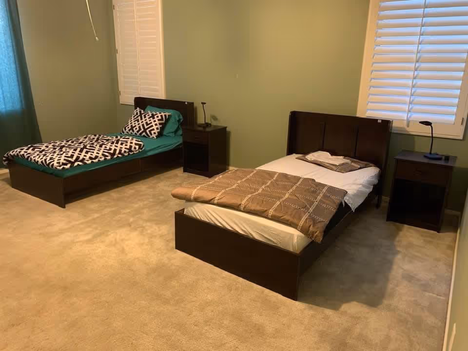 A bedroom with two single beds, each with a nightstand and a small lamp. The walls are painted green, and there are windows with white shutters. The floor is carpeted in beige.