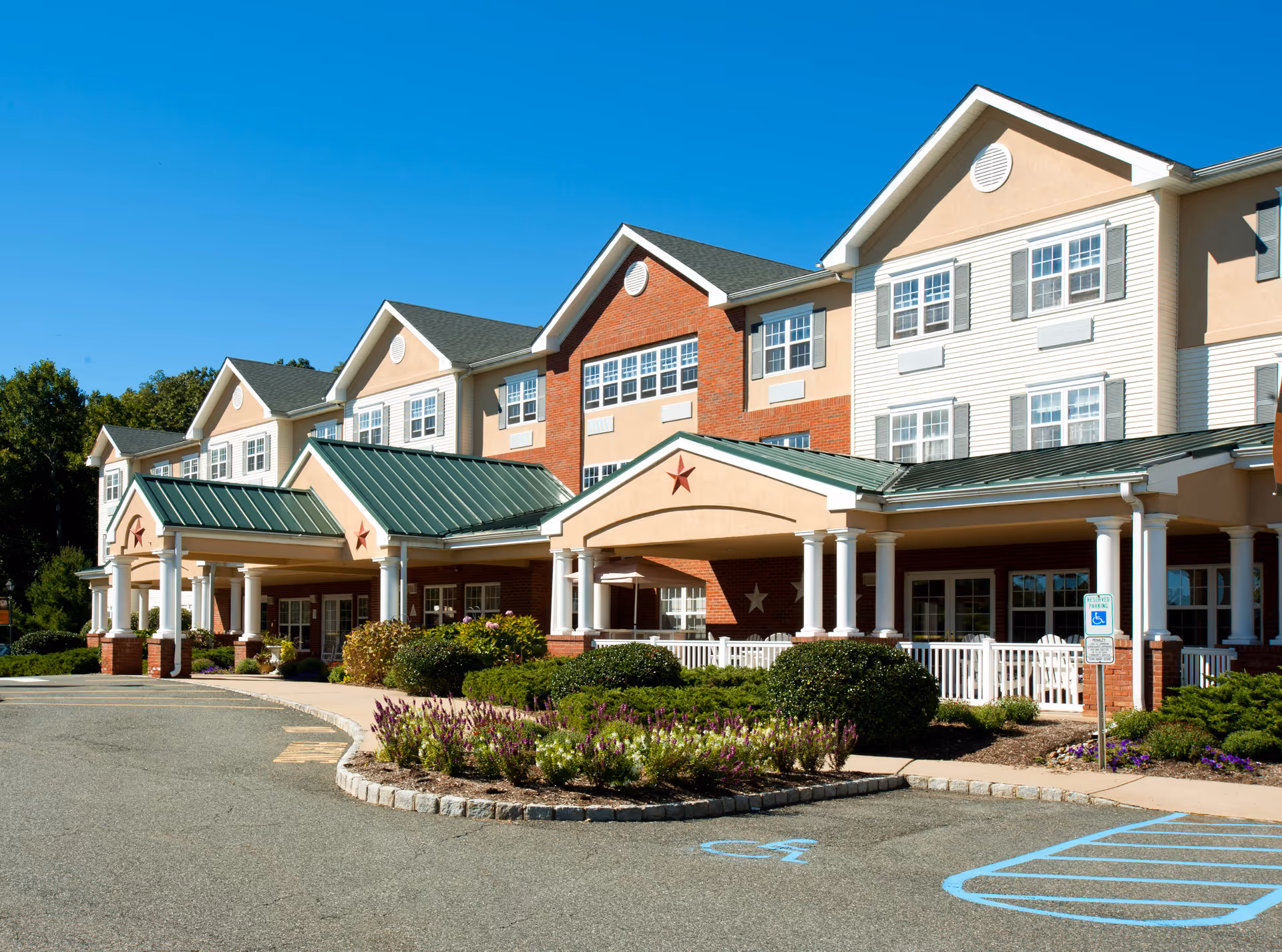 Exterior view of Mt. Arlington Senior Living facility showing a multi-story building with a green metal roof, beige and red brick walls, white columns, and a covered entrance. There are landscaped bushes and flowers in front, a clear blue sky, and a handicapped parking space visible in the foreground.