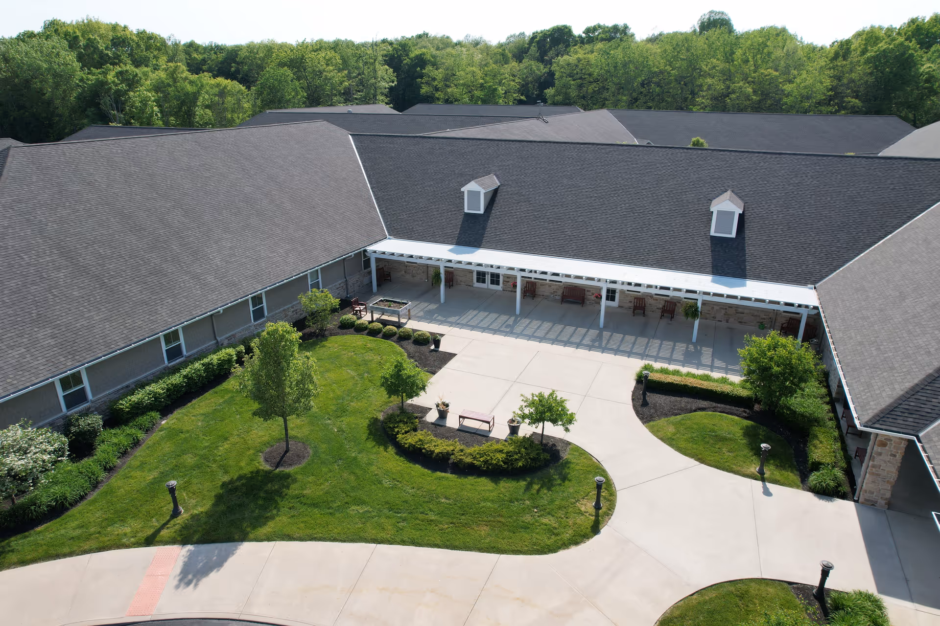 Aerial view of a senior living facility courtyard with well-maintained green lawns, small trees, shrubs, and a paved walkway. The building has a dark shingled roof and a covered porch area with benches and chairs. The surrounding area is lush with dense green trees.