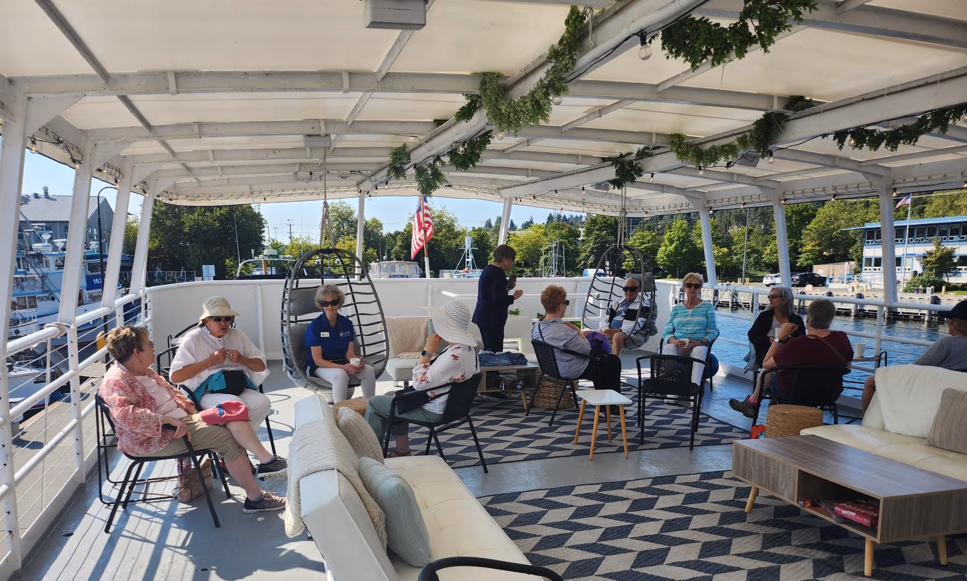 A group of elderly people sitting and chatting on a covered outdoor deck with comfortable seating, including chairs and sofas. The deck overlooks a marina with boats and trees in the background. The area is decorated with hanging greenery and string lights.
