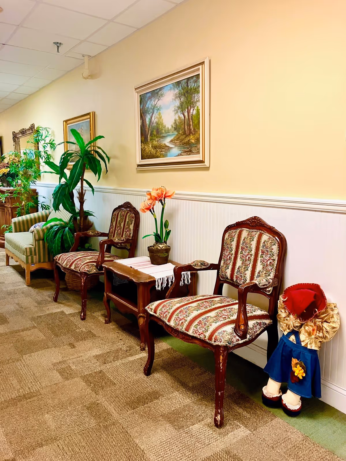 A hallway in a senior living facility with vintage-style upholstered chairs, a small wooden table with a potted flower, and framed landscape paintings on the wall. There are also several green plants and a decorative doll wearing a red hat and blue dress placed against the wall.