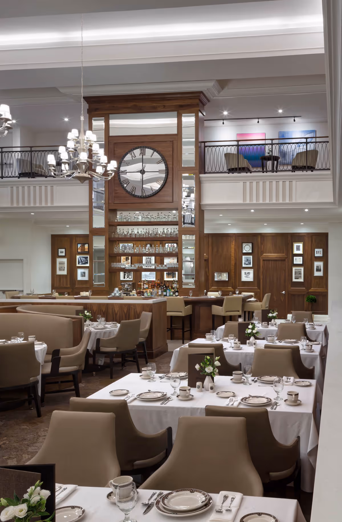 Elegant dining room with white-clothed tables and place settings, a central wood bar topped by a large clock, and a mezzanine seating area.