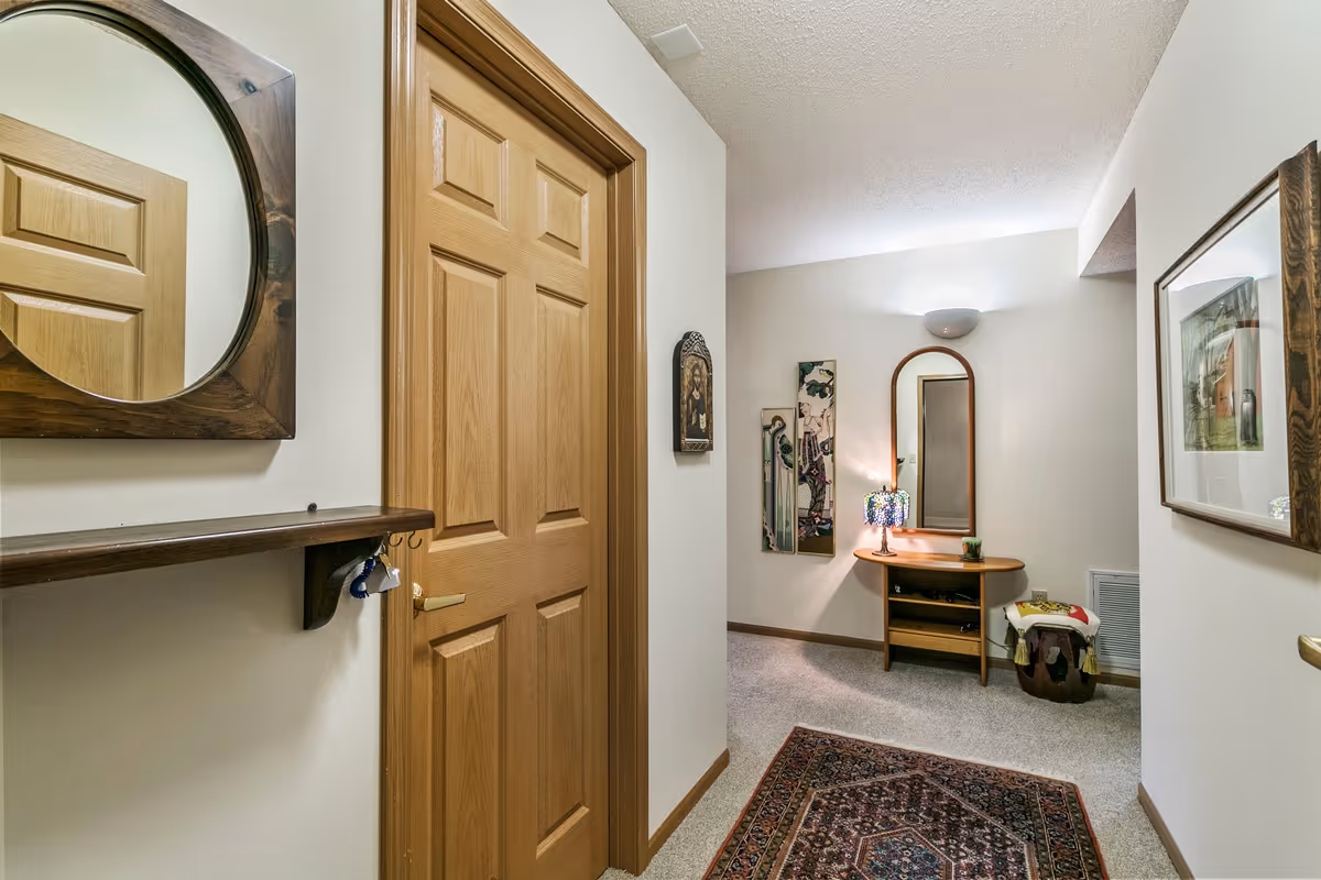 A hallway in a senior living facility with beige walls and carpeted floor. There is a wooden door on the left, a round wooden-framed mirror above a small shelf with hooks, and a patterned rug on the floor. At the end of the hallway, there is a small wooden table with a curved mirror, a decorative lamp, and a small plant. The walls are decorated with framed artwork and a small religious icon.