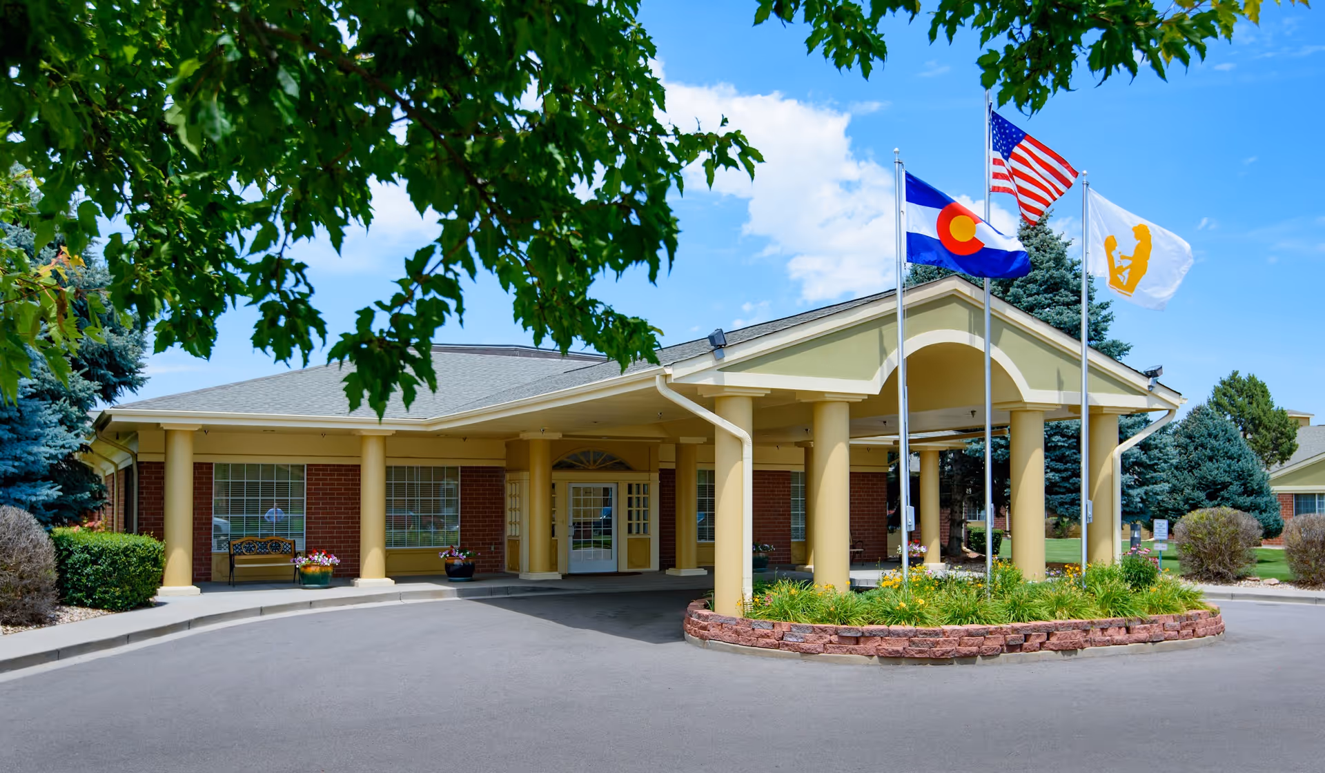 Front exterior view of Hallmark Nursing Center building with a covered entrance supported by columns. Three flagpoles display the Colorado state flag, the American flag, and a white flag with a yellow emblem. There are trees and shrubs around the building and a clear blue sky with some clouds.