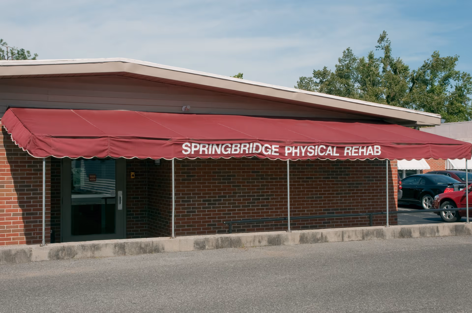 Exterior view of a brick building with a maroon awning that reads 'SPRINGBRIDGE PHYSICAL REHAB'. There is a glass door entrance under the awning and a parking lot with cars visible to the right.